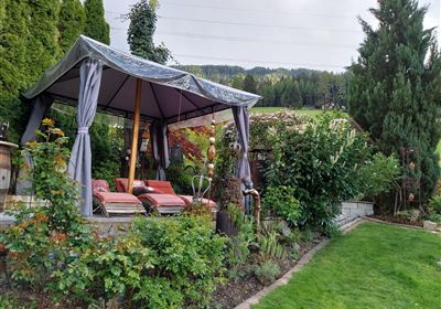 A cozy pavilion in the garden, surrounded by green plants and shrubs. The seating areas invite you to relax.